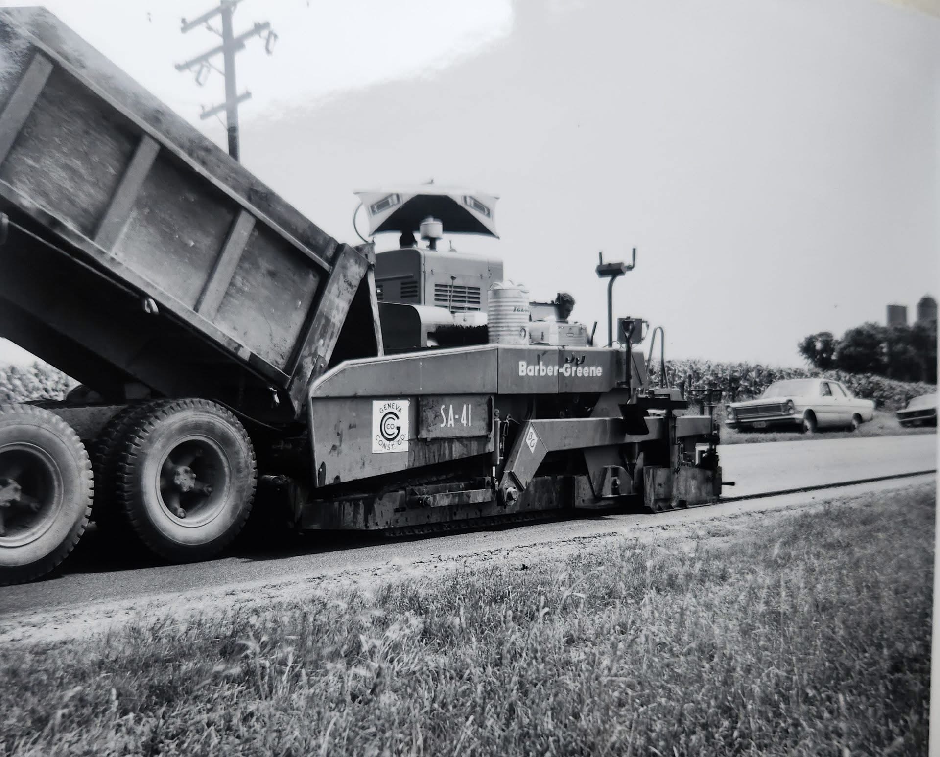 A Barber-Greene SA-41 asphalt paver, unit marked and decalled GENEVA CONST. CO, laying a mat on a rural two-lane road while a tandem dump truck feeds its hopper. A corn field and a farm silo stand behind it; a mid-1960s car passes on the finished lane.